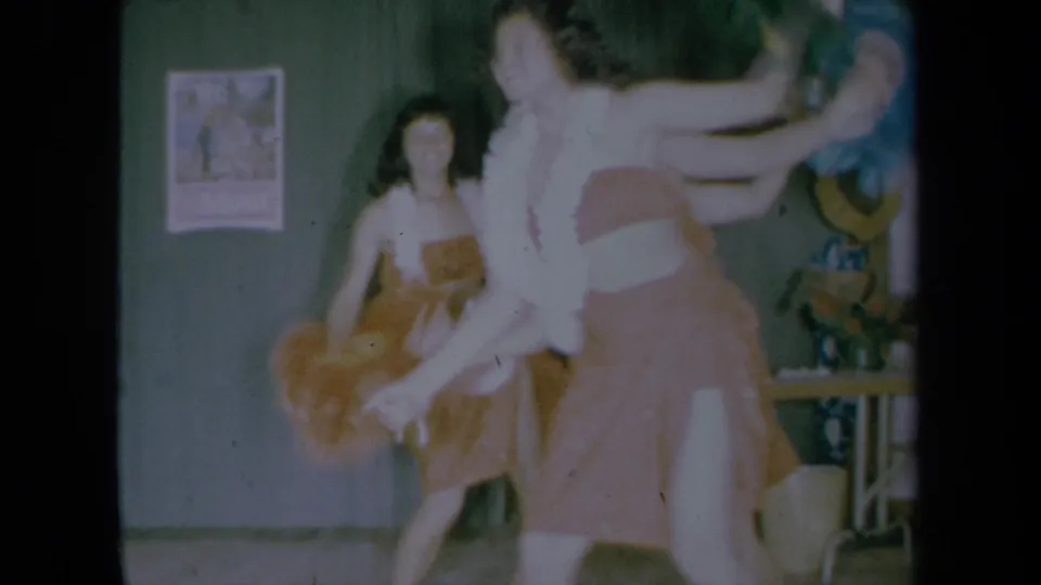 Two young women dance in a California home interior, one in a pink dress with a white feather boa and another in a brown dress.