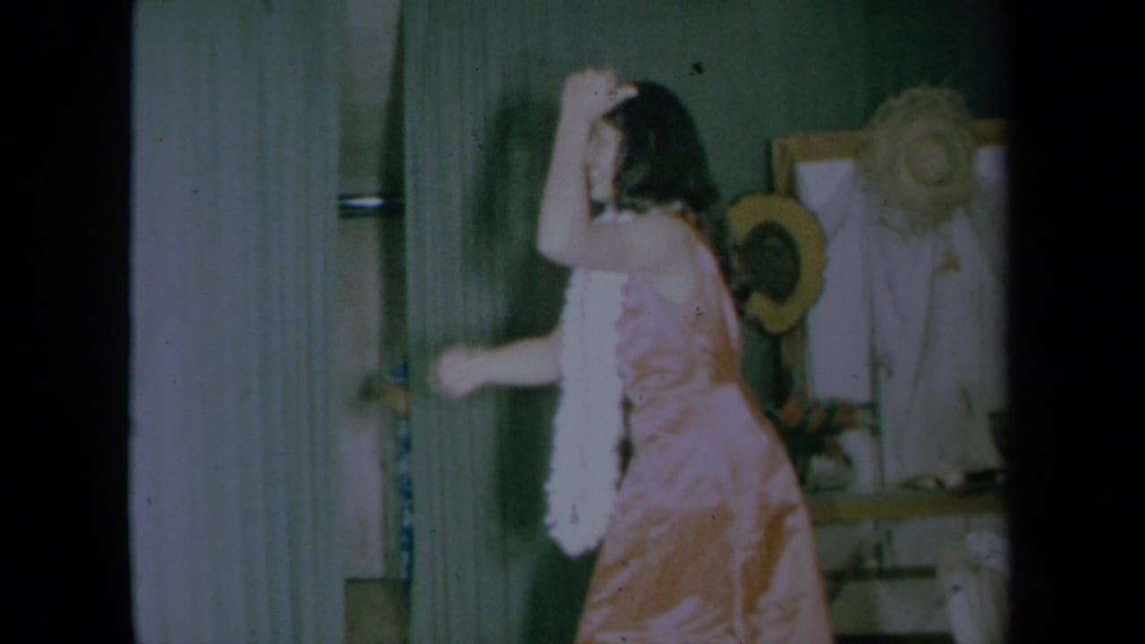 A young woman in a pink dress and white boa dances beside a mirror and hanging hat.