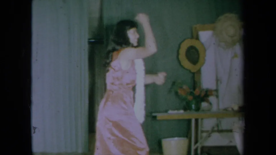 A young woman in a pink satin-style dress and white feather boa dances in front of a green wall in the California home-movie scene.