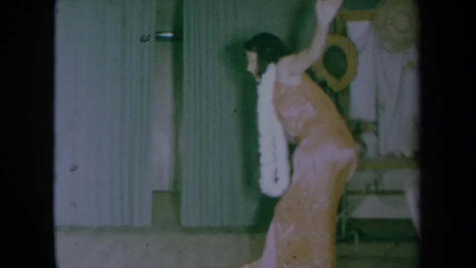 A young woman in a pink dress with a white feather boa leans into a dance move near a mirror, flowers, and a straw hat.