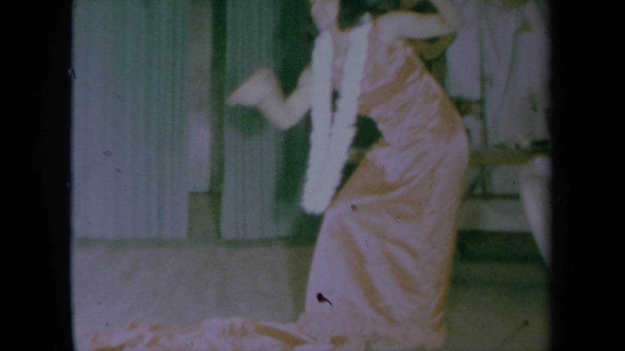 In 1961 California home-movie footage, a woman in a pink satin dress and white lei dances indoors near green curtains.