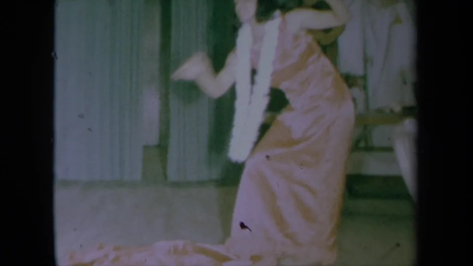 In 1961 California home-movie footage, a woman in a pink satin dress and white lei dances indoors near green curtains.