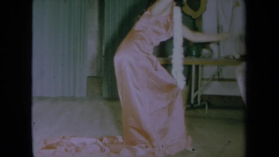 The woman in a pink dress and white lei leans into a dance gesture inside a California home setting from 1961.