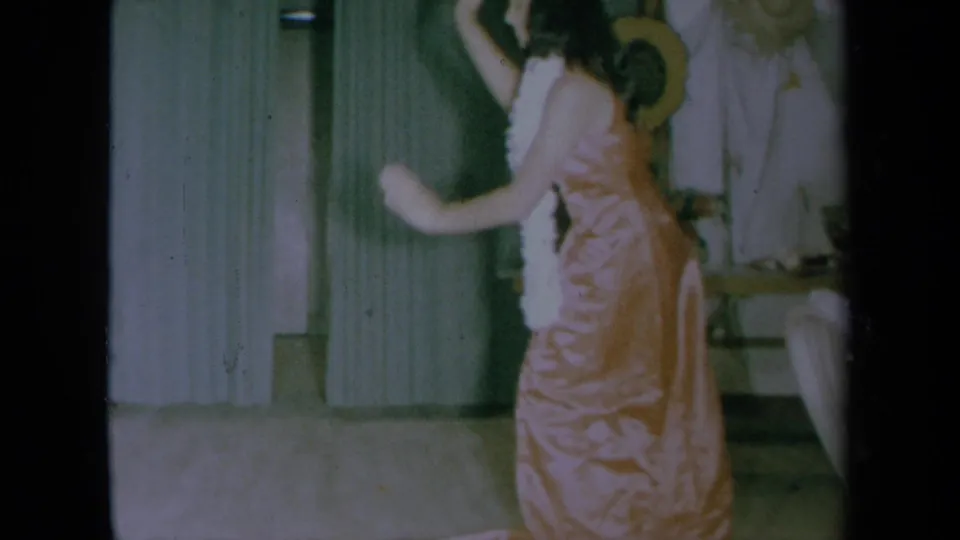 A side view shows the woman moving her arms while dancing in a pink dress and lei in the 1961 indoor home-movie scene.