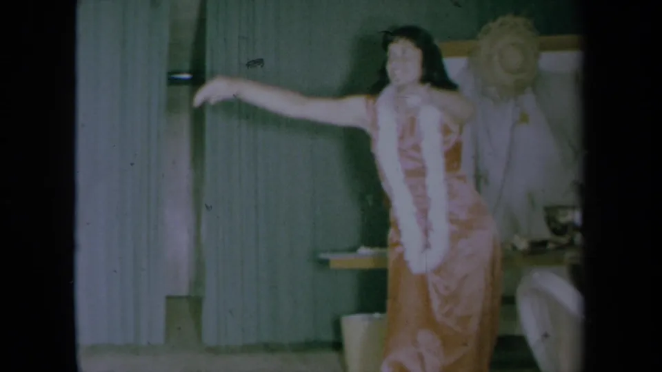 The woman faces the camera with one arm extended, wearing a pink dress and white lei in a 1961 California home interior.