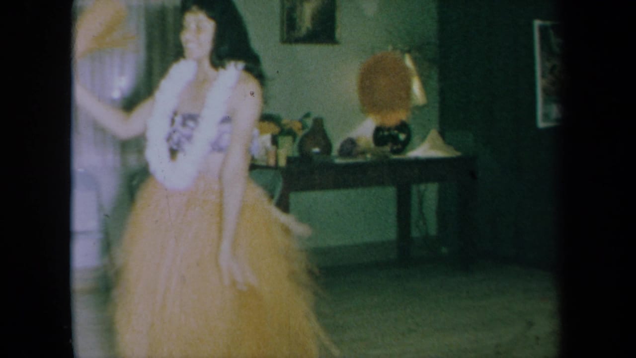 In 1961 California home-movie footage, a young woman dances indoors in a grass skirt and white lei near curtains, wall art, and a lamp.