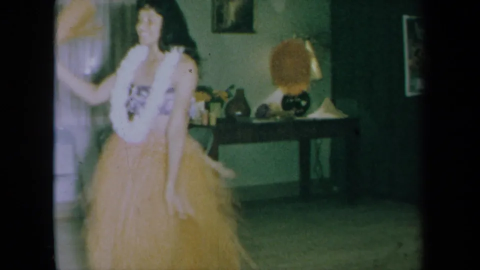 In 1961 California home-movie footage, a young woman dances indoors in a grass skirt and white lei near curtains, wall art, and a lamp.
