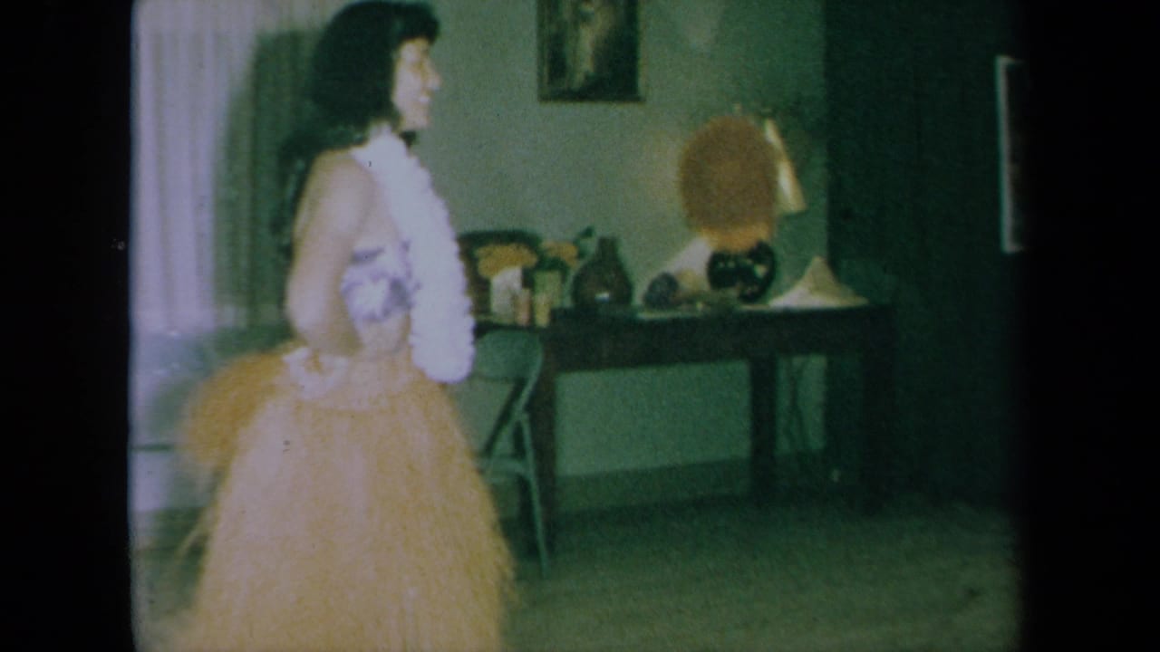 Young woman in hula costume turns sideways in a living room with a lamp and wall pictures.