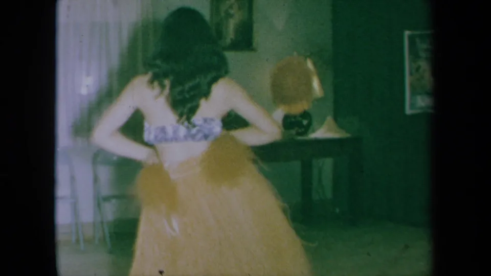 A rear view shows the dancer in a grass skirt and patterned top, with a folding chair, curtains, and table lamp in the living room.