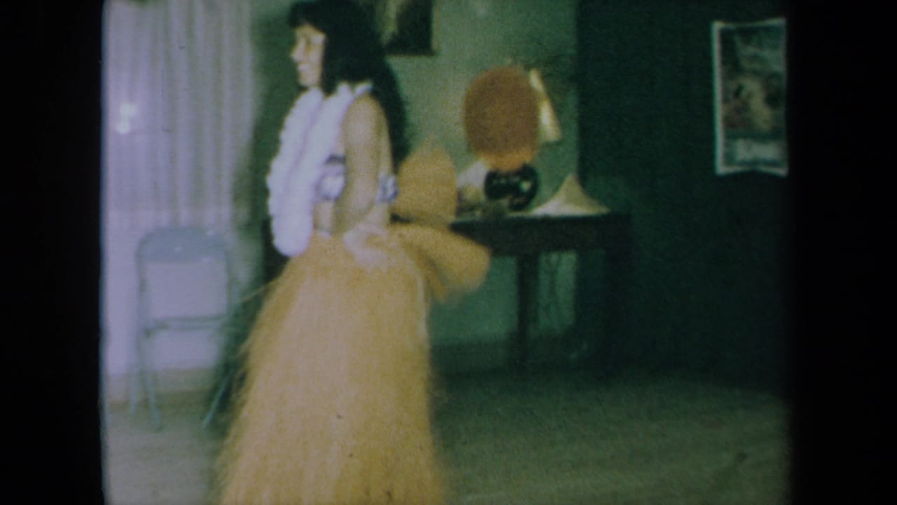 In a 1961 California home-movie frame, an adult woman in a grass skirt and white lei dances in a dim private room.