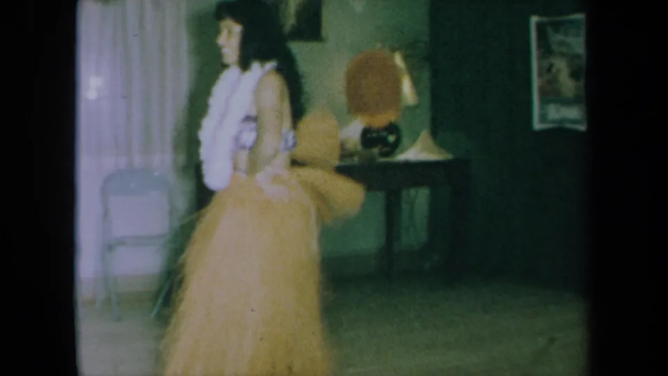 In a 1961 California home-movie frame, an adult woman in a grass skirt and white lei dances in a dim private room.