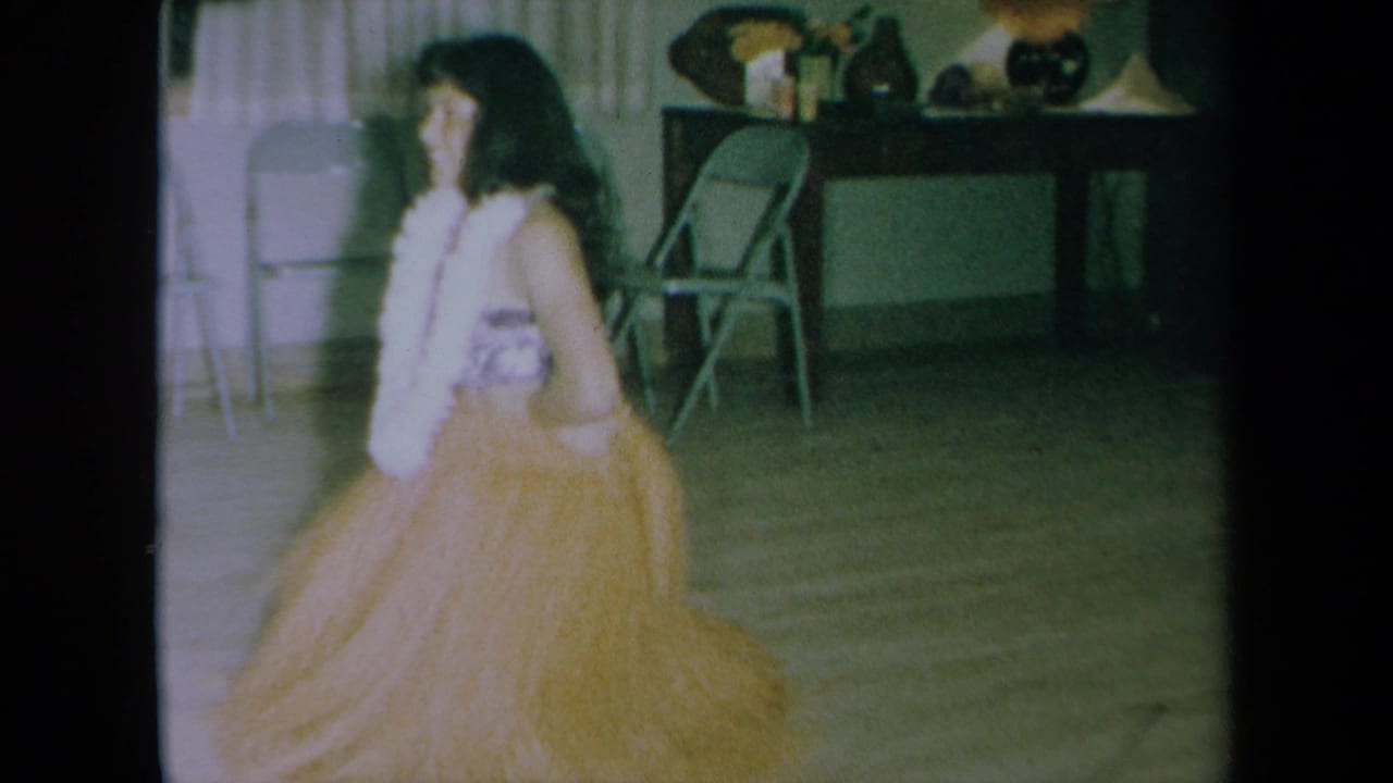 Woman in Hawaiian-style costume turns beside curtains, folding chairs, and a table with decor.
