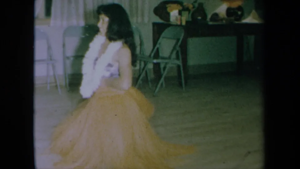 The dancer continues her routine in the dim 1961 California home-movie interior, with chairs and table decor visible.