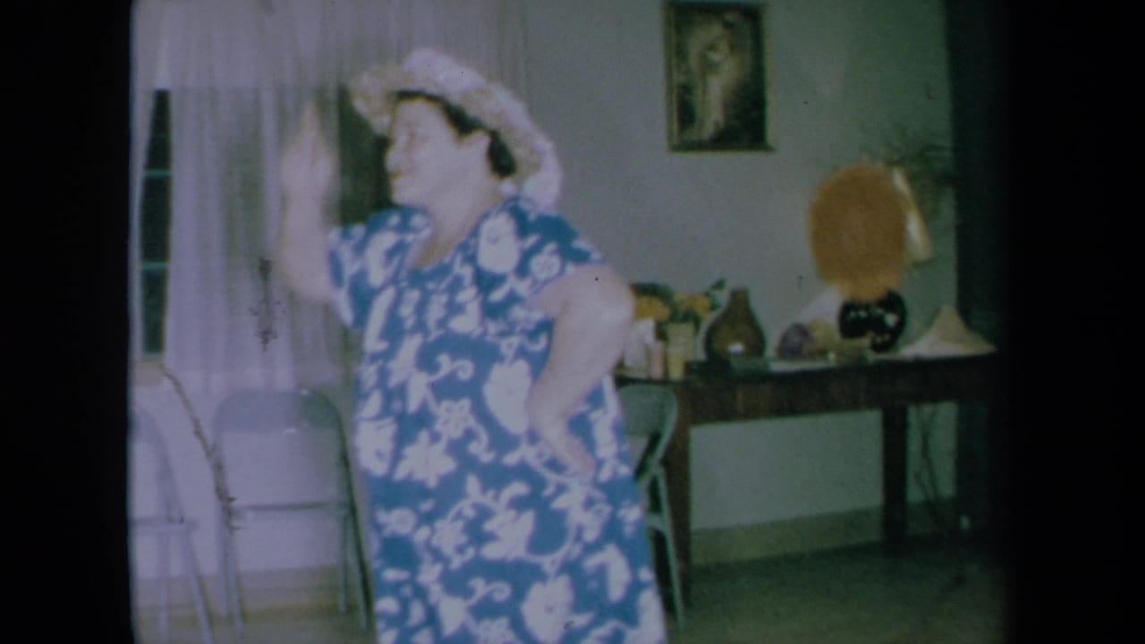Woman in a blue floral dress and tan hat dancing in a home interior.
