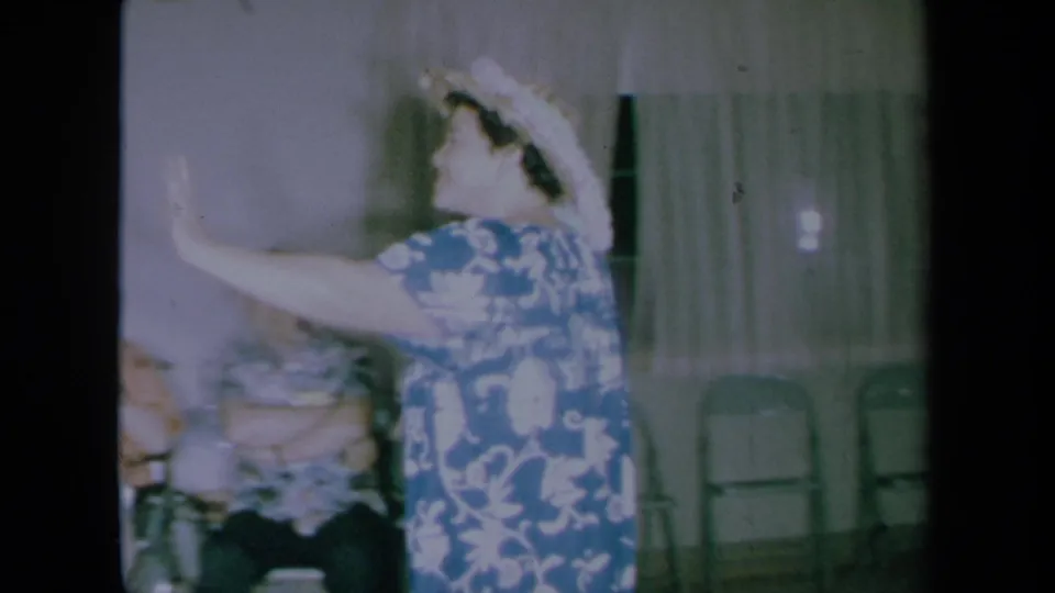 The woman in the blue floral dress gestures mid-dance in a private California home, with seated adults, curtains, and folding chairs behind her.