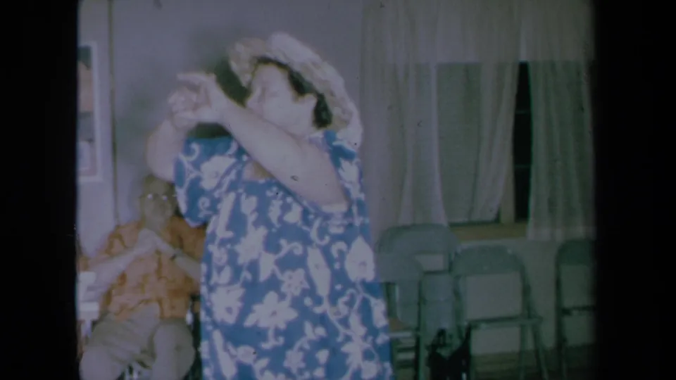 A 1961 home-movie frame shows the woman raising her arms in dance, with a seated adult, curtained windows, and folding chairs in the room.