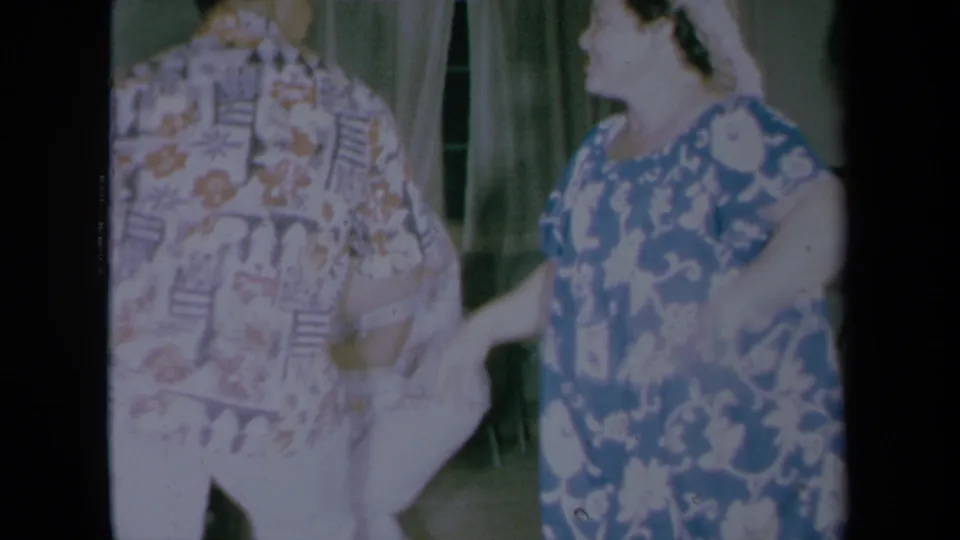 The woman continues dancing beside a man in a patterned shirt inside the private home, with curtains and folding chairs visible in the background.