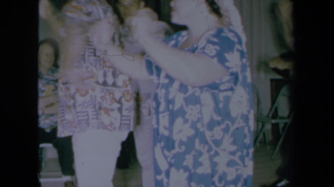 1961 California home-movie frame showing a woman in a blue floral muumuu dancing beside guests in aloha shirts inside a private room.