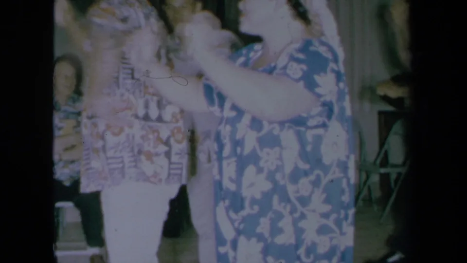 1961 California home-movie frame showing a woman in a blue floral muumuu dancing beside guests in aloha shirts inside a private room.