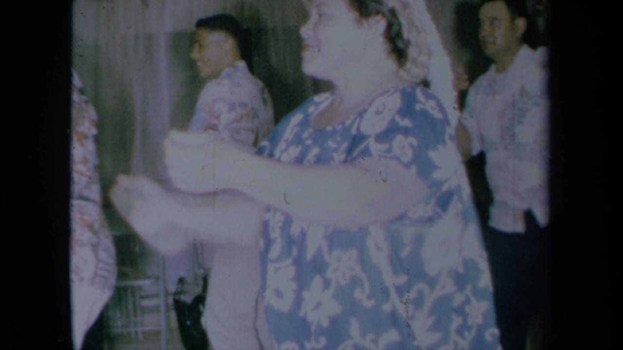 Dancing woman in floral dress moves her arms as men in aloha shirts stand nearby.
