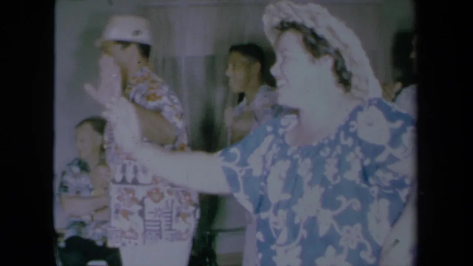 1961 California 8mm frame of a smiling woman dancing in a floral muumuu, with seated and standing guests visible behind her.