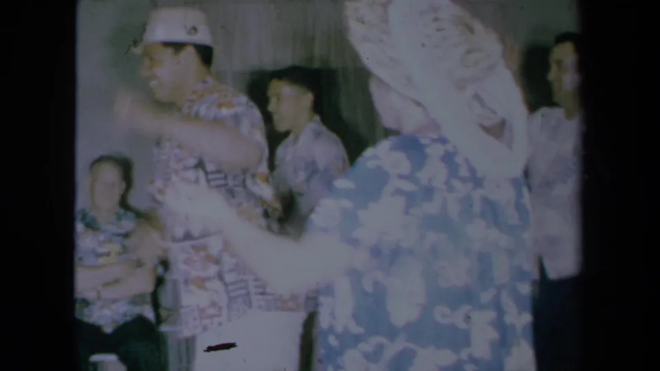 A woman in a blue floral muumuu turns toward a man in an aloha shirt as other guests watch in this 1961 California home-movie frame.