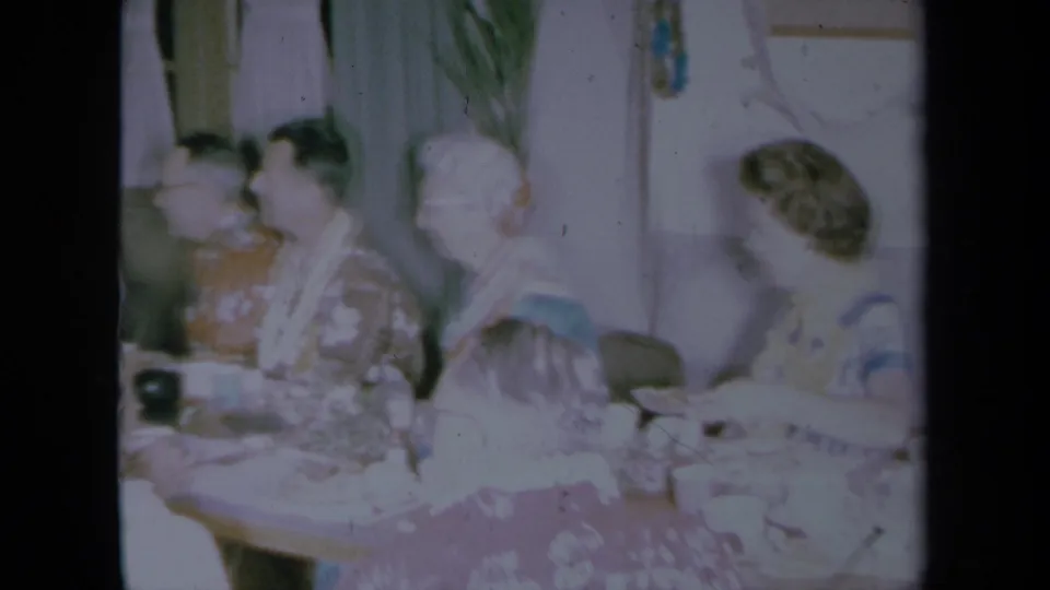 1961 California 8mm frame showing older adults in floral clothing gathered around a table in a softly lit interior.
