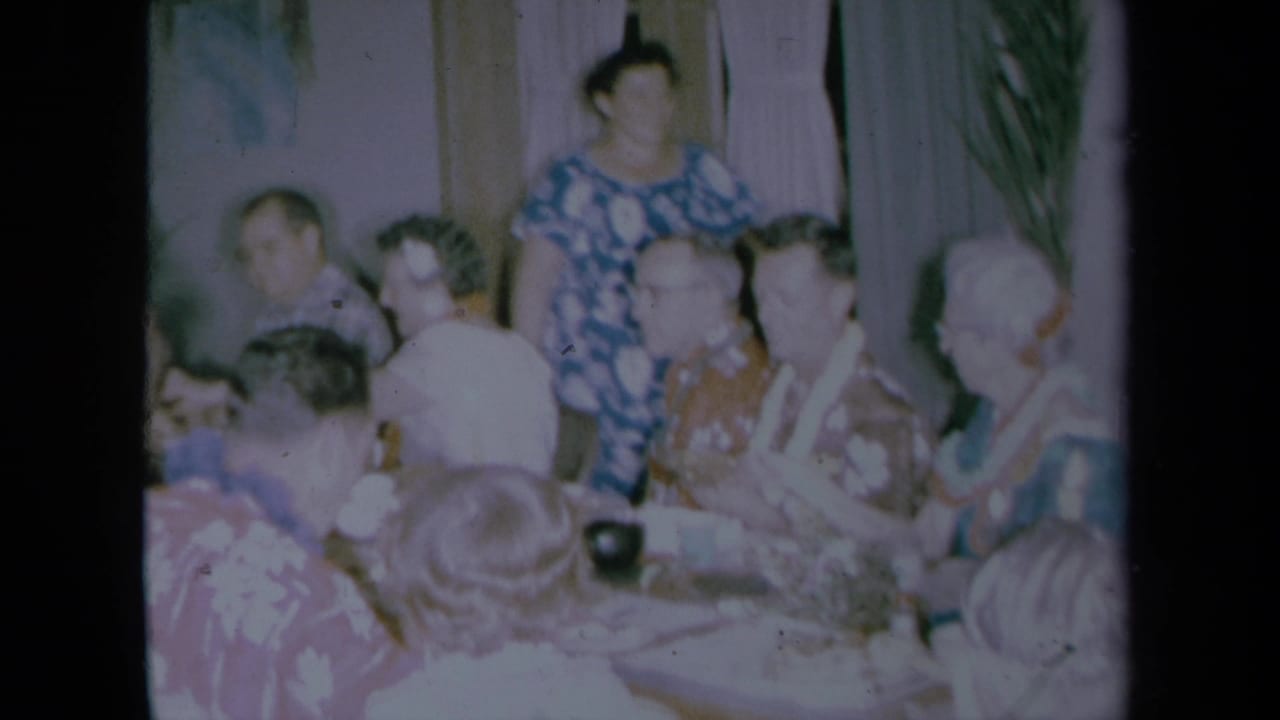 1961 California home-movie frame with seated older adults in patterned shirts and leis, plus a standing woman in the background.
