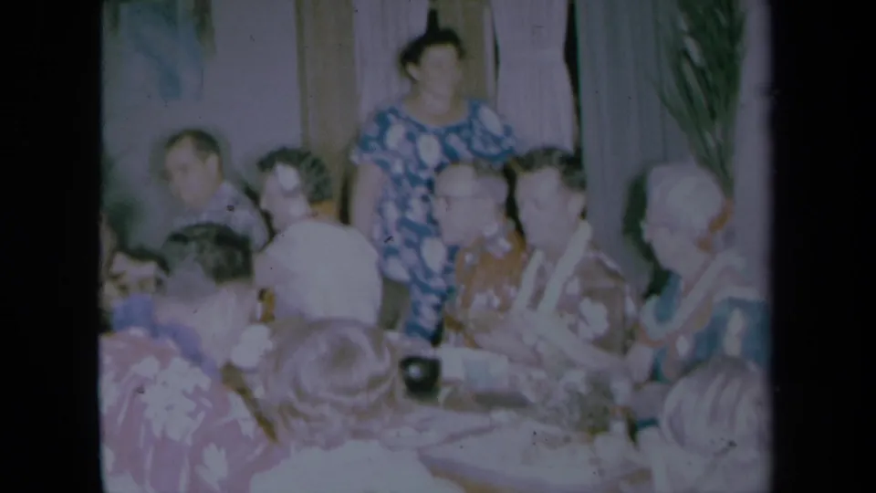 1961 California home-movie frame with seated older adults in patterned shirts and leis, plus a standing woman in the background.