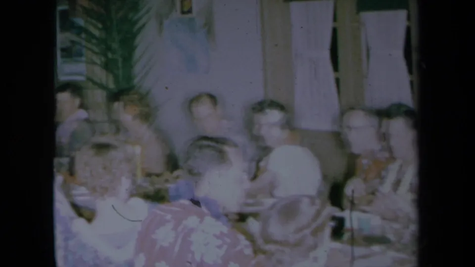 Soft 8mm frame from a 1961 California interior showing adults gathered at a table, with motion blur and floral clothing visible.