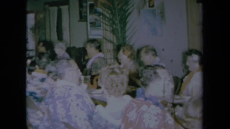 1961 California home-movie frame showing a crowded indoor table with older adults in floral shirts and leis near tall houseplants.