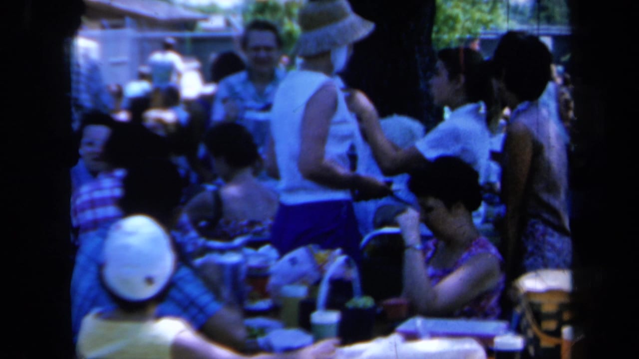 People stand and sit near a table while a woman in a straw hat serves food.
