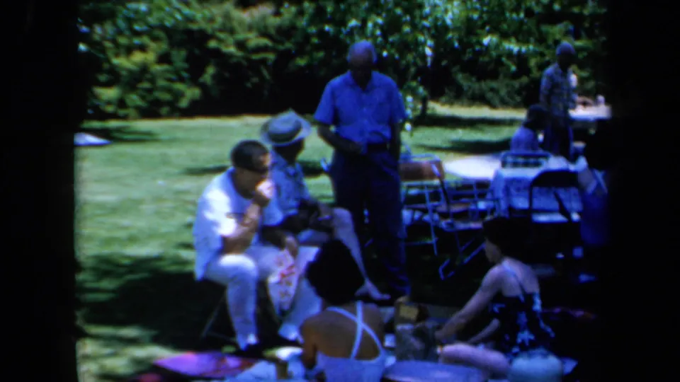 1961 California home-movie frame showing people seated on a lawn beside folding chairs and picnic tables.