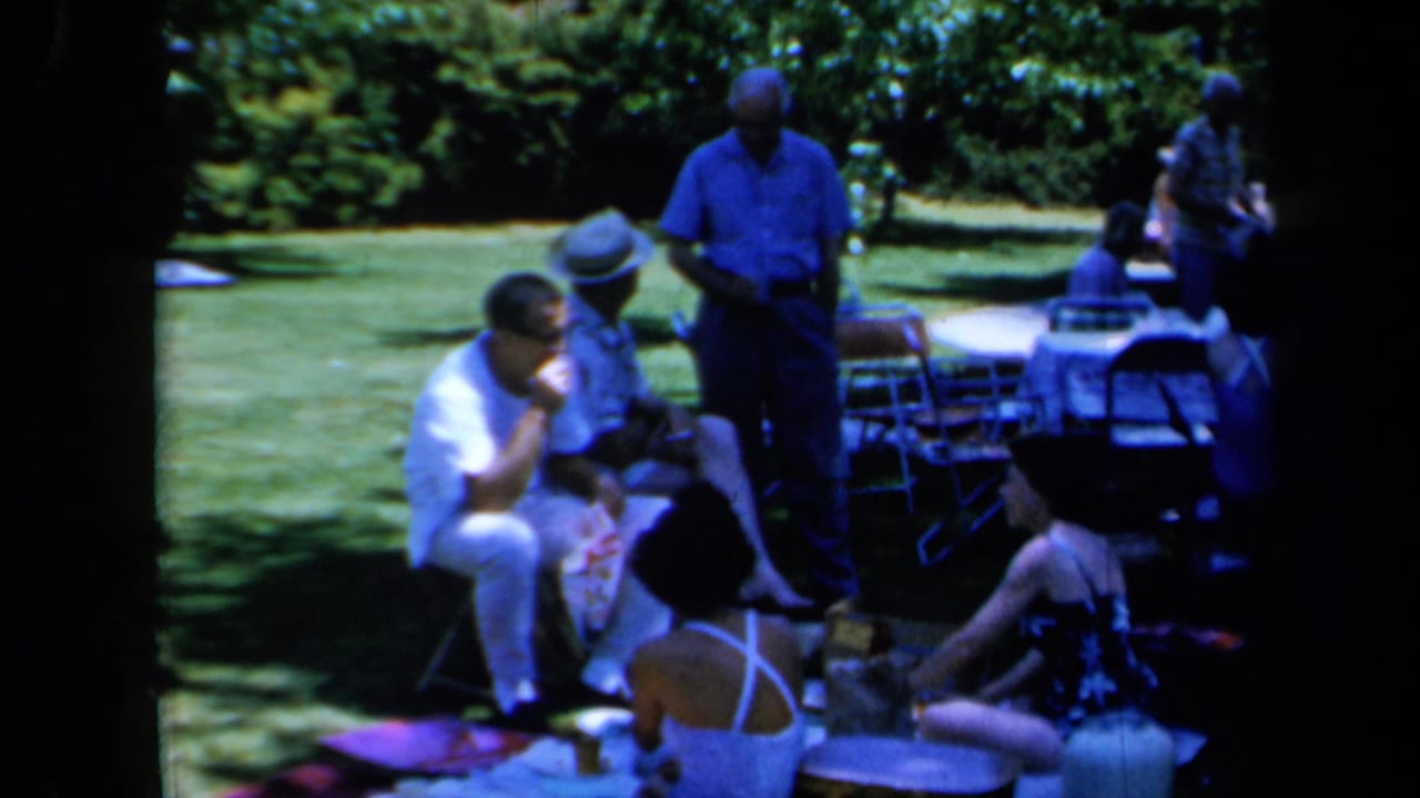 1961 California home-movie frame showing adults in summer clothing seated on blankets and chairs beside picnic items on a shaded lawn.