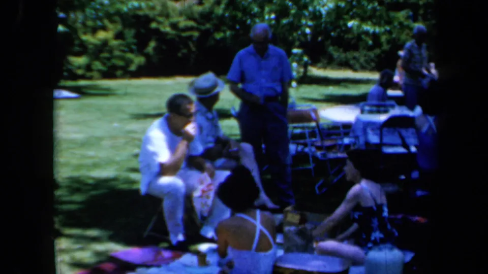 1961 California home-movie frame showing adults in summer clothing seated on blankets and chairs beside picnic items on a shaded lawn.