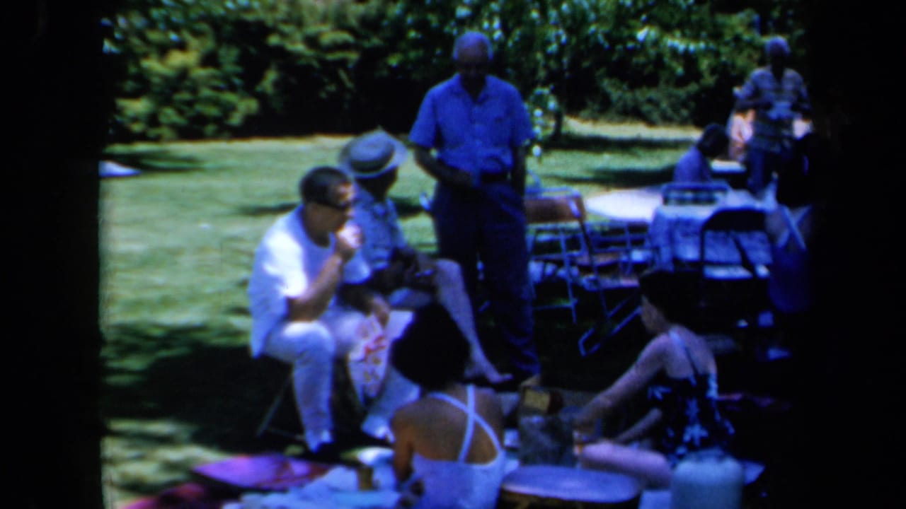 Adults gather on a lawn with chairs, blankets and a picnic table nearby.