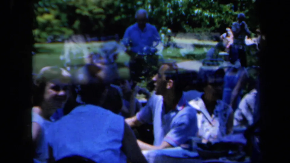 Handheld 8mm frame of adults seated close together at a 1961 California picnic, with faces, shirts and shaded greenery visible.