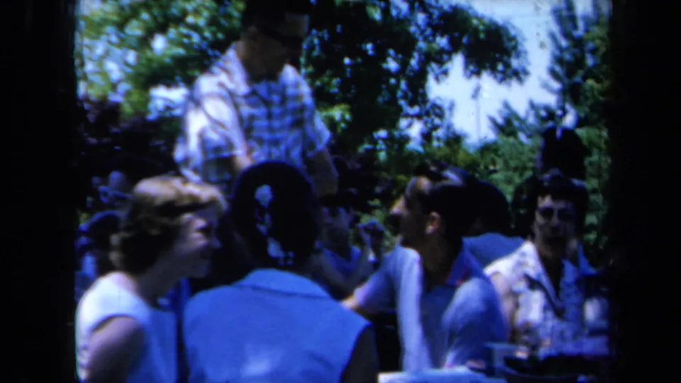 Outdoor 1961 California picnic scene with several adults seated in conversation and one person standing nearby under trees.