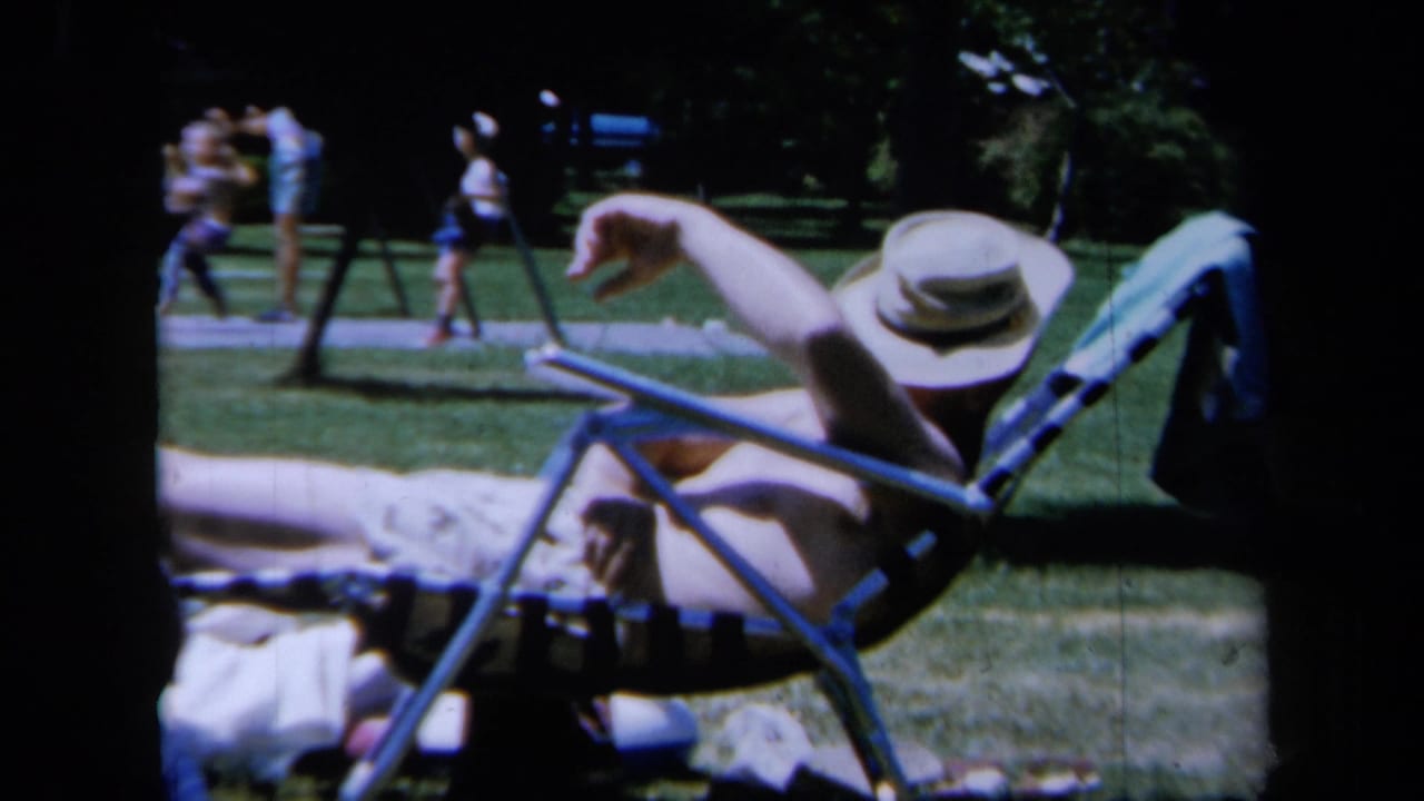 1961 California park home-movie frame showing a person sunning on a striped lounge chair with blankets and distant visitors on the grass.