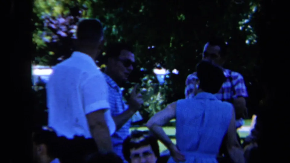 1961 California park frame showing adults in casual summer clothing gathered in conversation, with visible faces and shaded greenery.