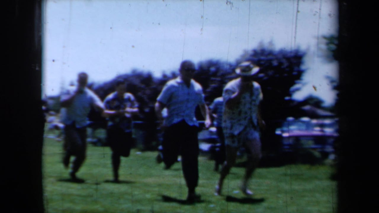 In 1961 California home-movie footage, adults run toward the camera on a sunny lawn during a casual athletic challenge.
