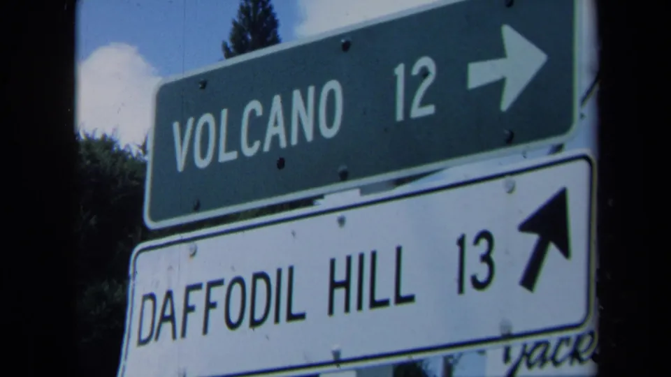 1969 home-movie frame showing weathered road signs for Volcano and Daffodil Hill, captured with visible 8mm grain and slight motion softness.