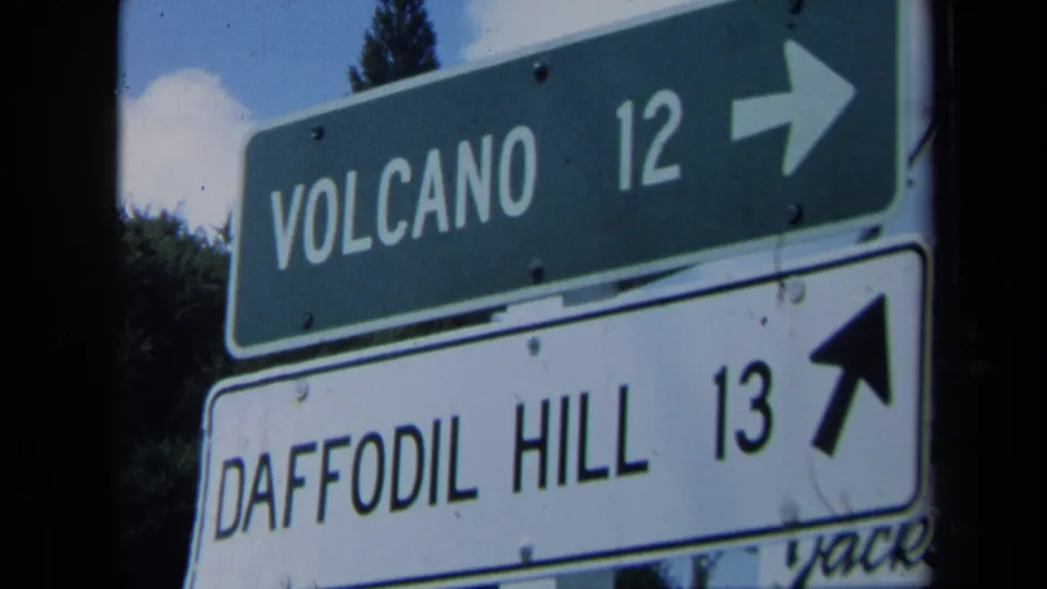 Final 8mm frame of the California roadside signs, with Volcano and Daffodil Hill directions partly softened by film grain and camera movement.