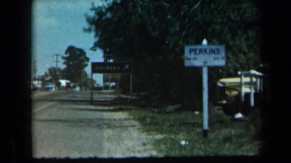 Wider 1969 California roadside view with Jackson and Perkins signs, trees, utility poles, and cars in the distance.