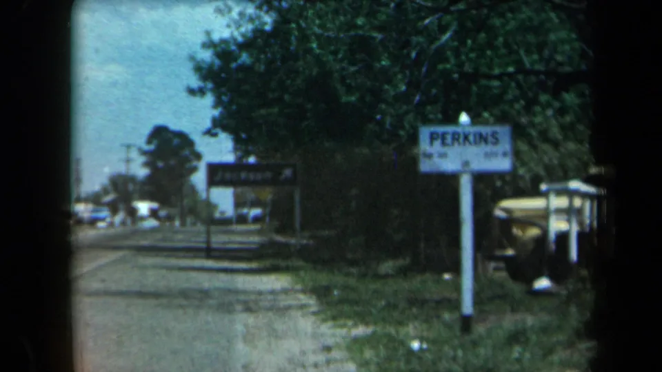 California 8mm frame of a rural roadside with Perkins and Jackson signs, roadside trees, and vintage cars passing farther down the road.