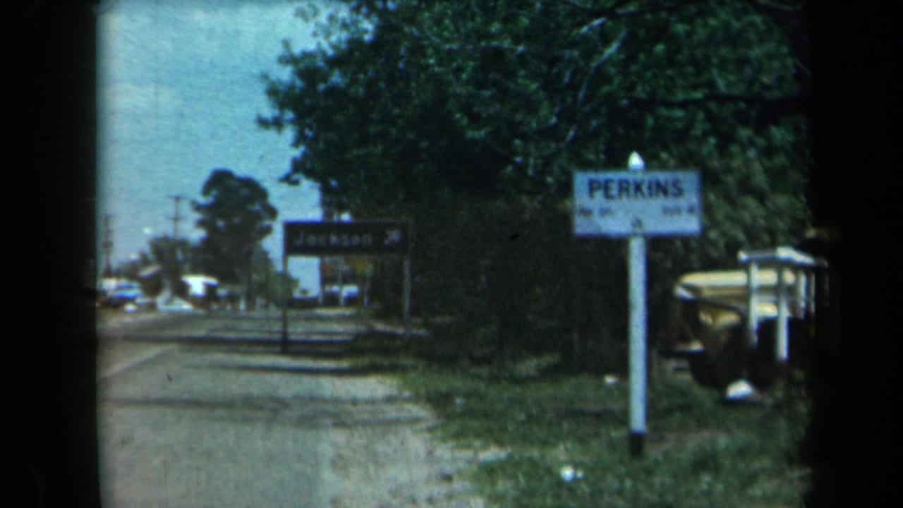 1969 California 8mm roadside frame showing a Perkins sign, distant Jackson direction sign, trees, utility poles, and a vehicle near the shoulder.