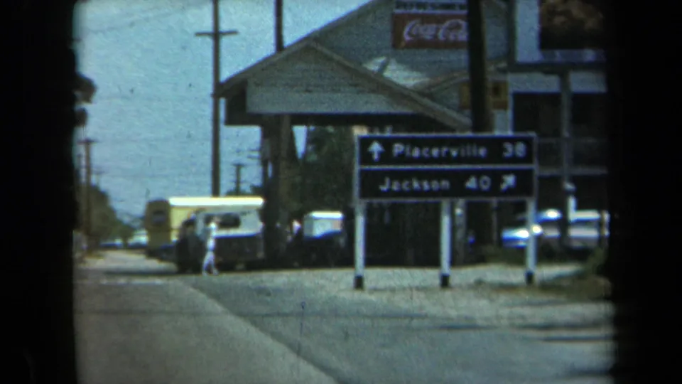 1969 California roadside scene with Placerville and Jackson sign, white van, parked cars, Coca-Cola sign, and a person by the building.