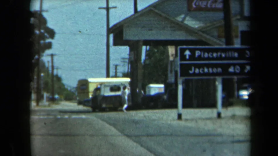 1969 California 8mm frame showing the Placerville and Jackson direction sign beside a white van, parked cars, building frontage, and road edge.