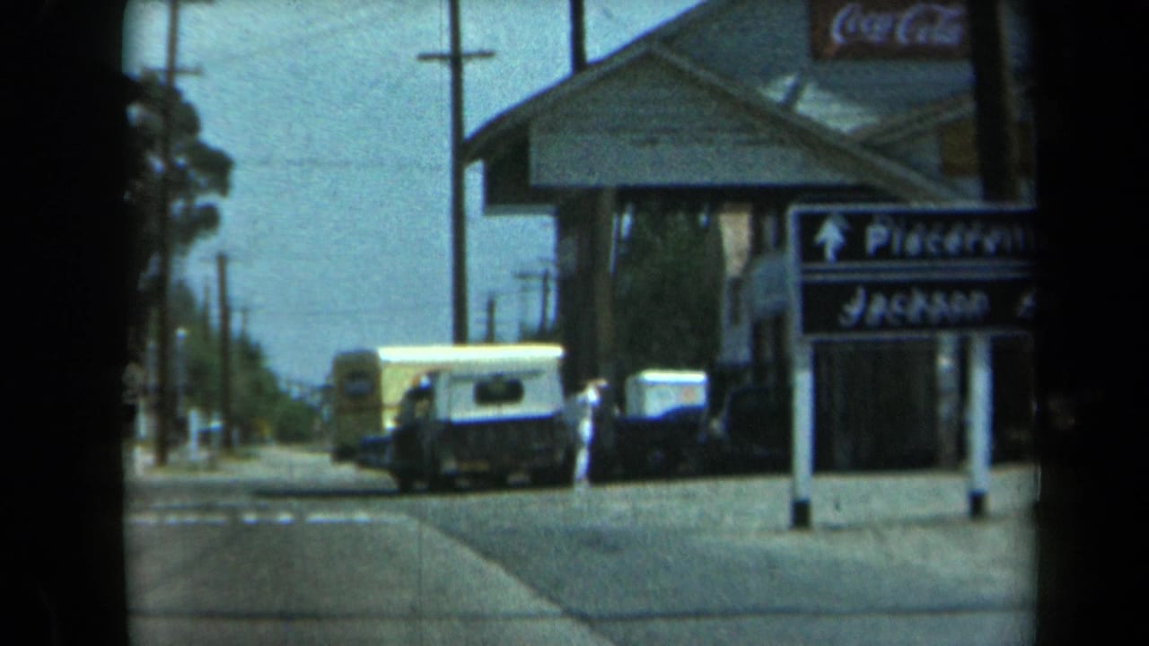 1969 California 8mm frame showing a small roadside store, parked vans, utility poles, trees, and a person walking near the storefront.