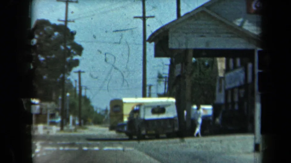 1969 California street view with a roadside storefront, parked van, utility poles, trees, and a person in light clothing near the entrance.